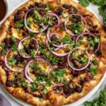 Crispy Oven-Baked Birria Pizza topped with melted cheese, shredded beef birria, red onion, and fresh cilantro, served with consommé and lime wedges
