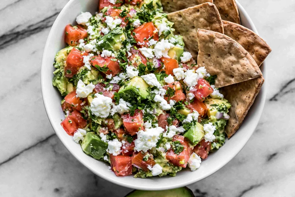 Avocado Feta Dip topped with crumbled feta, diced tomatoes, fresh parsley, and creamy avocado, served with crispy crackers.