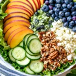 Peach Berry Summer Salad featuring sliced peaches, fresh blueberries, cucumber rounds, feta cheese, chopped pecans, and leafy greens with lemon basil dressing.