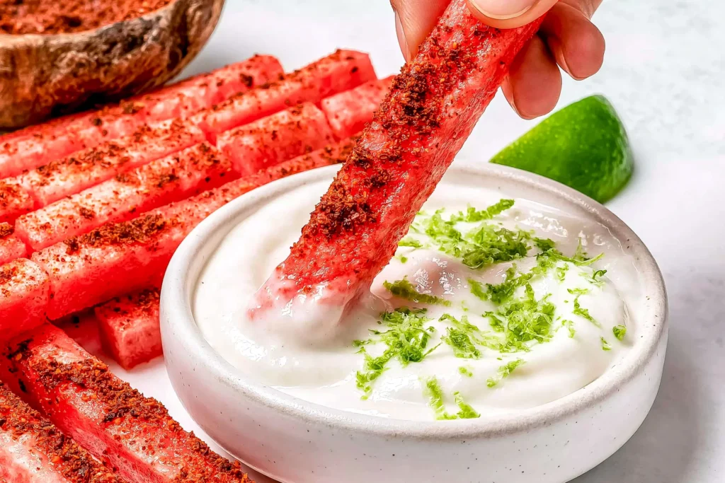 Hand dipping Spicy Watermelon Fries coated in chili lime seasoning into a creamy lime yogurt dipping sauce topped with fresh lime zest.