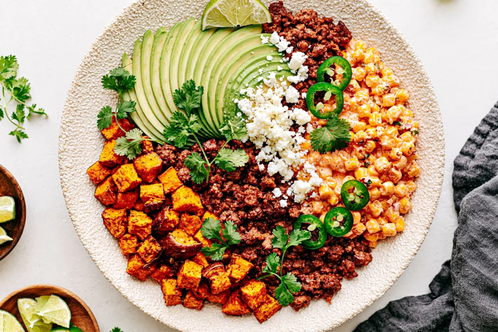 Street Corn Beef Bowl topped with seasoned ground beef, roasted sweet potatoes, creamy Mexican street corn, sliced avocado, cotija cheese, jalapeños, cilantro, and fresh lime.